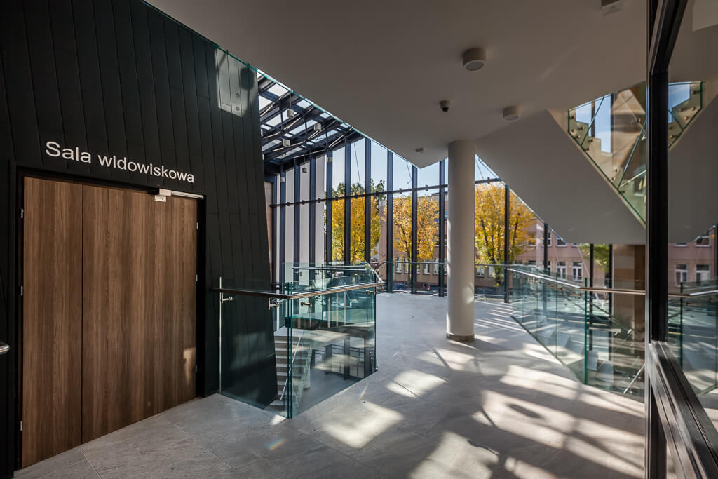 Wejście do sali widowiskowej, w oddali inne budynki widoczne przez przeszkloną ścianę budynku / Entrance to the auditorium, in the distance other buildings visible through the glass wall of the building