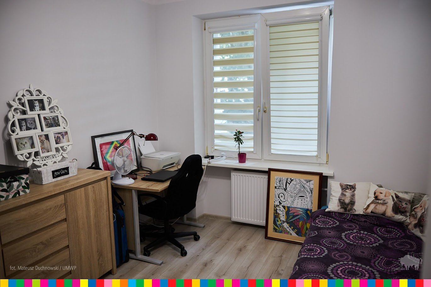 Wnętrze pokoju, po lewej stronie stoi biurko i krzesło. Na biurku stoją laptop, lampka, grafika w ramce. Po prawej stronie stoi łóżko.