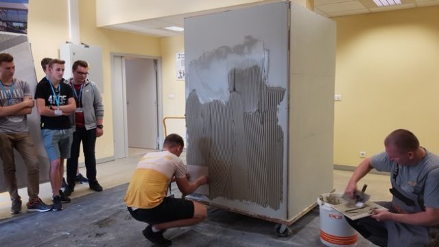 Grupa uczniów podczas wizyty edukacyjnej w przedsiębiorstwie patrzy na ucznia praktykującego szpachlowanie ściany. Po drugiej stronie pracownik firmy przygotowuje narzędzia do ćwiczeń. 